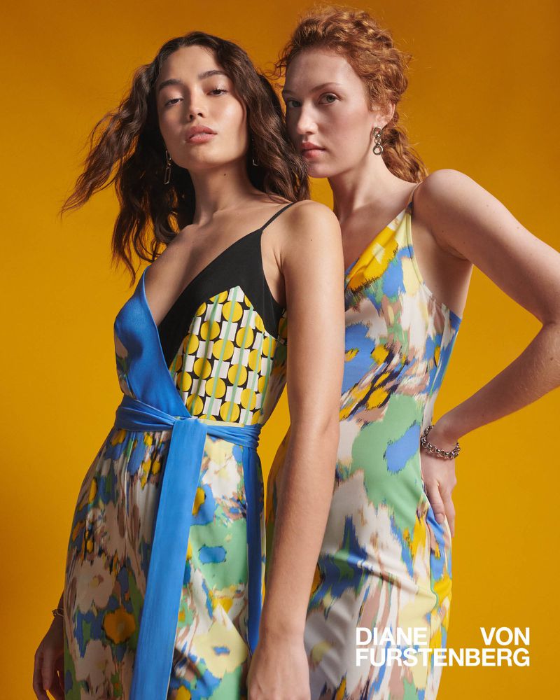 Two adult women in flowing sundresses stand against a yellow backdrop in a fashion shot with warm, diffused lighting.