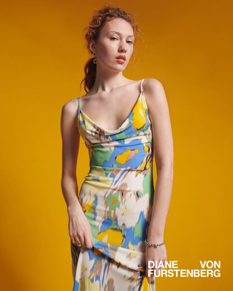 A woman in a printed dress stands against a yellow backdrop, showcasing a fashion-forward look with a focus on the dress's vibrant colors.