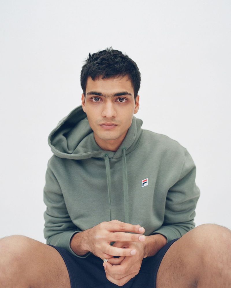 Man in olive green Fila hoodie looking at the camera, seated indoors against a white backdrop in soft, diffused studio lighting.
