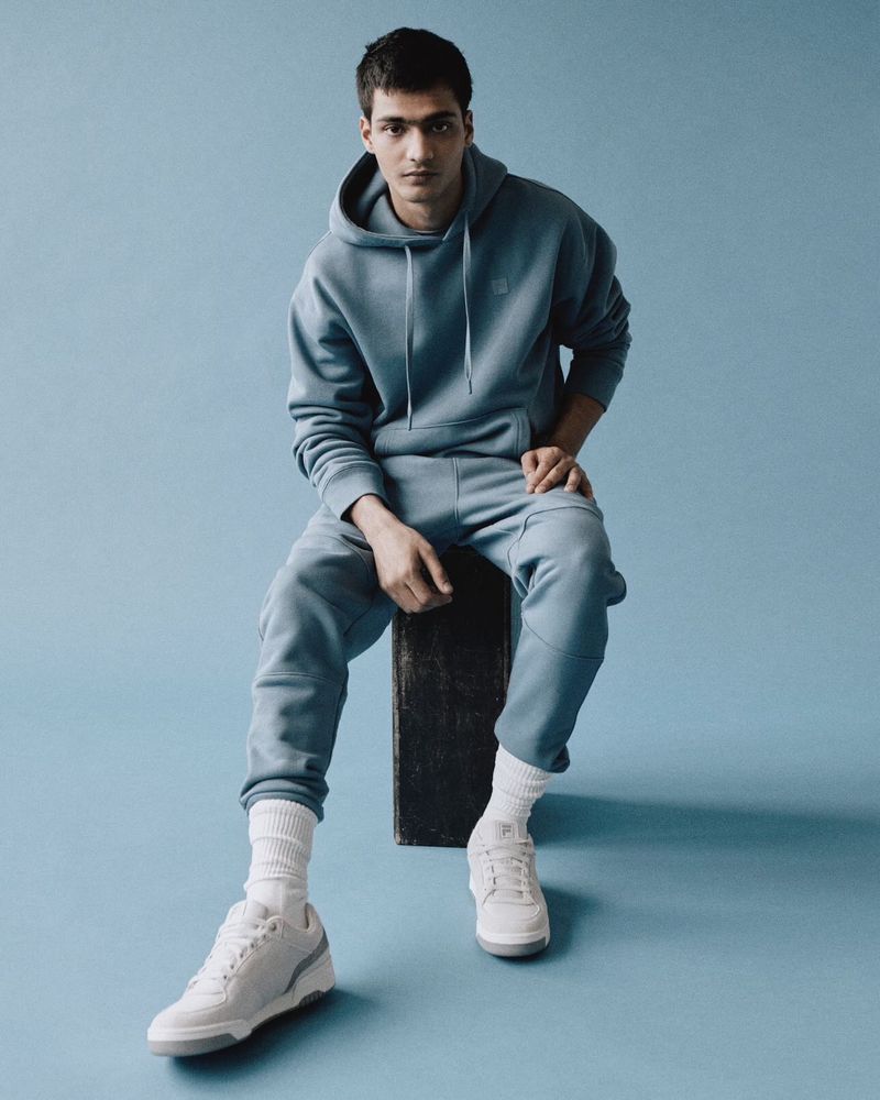 Portrait of a stylish young man in a blue sweatsuit sitting on a black block wearing white sneakers against a blue background.