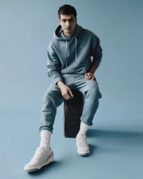 Man wearing a blue hoodie and sweatpants seated on a box against a blue studio background
