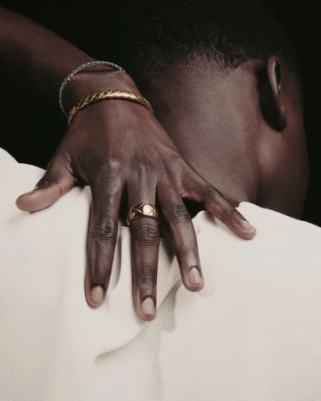 Close-up of a dark-skinned adult male's hand with gold rings and bracelets, resting on a person's shoulder against a dark backdrop.