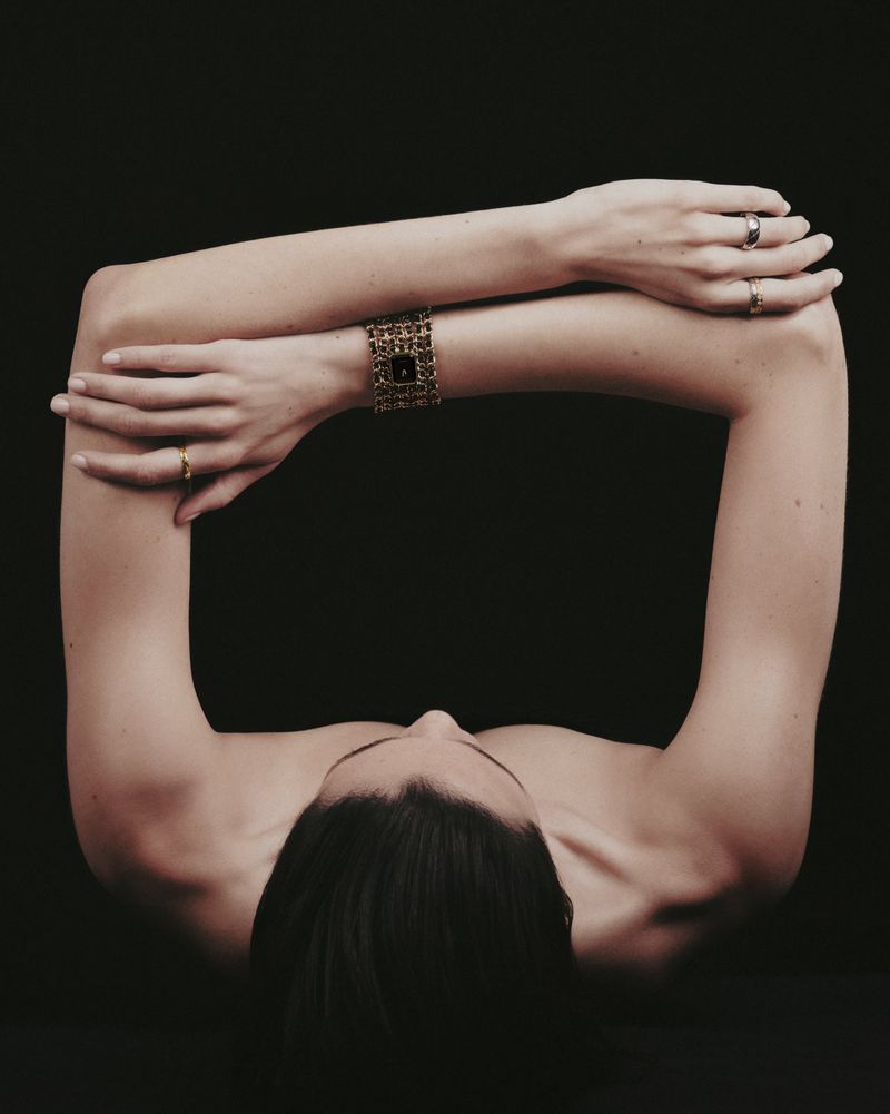 Overhead studio shot of a person with arms crossed, featuring a gold bracelet watch and rings against a solid black backdrop.