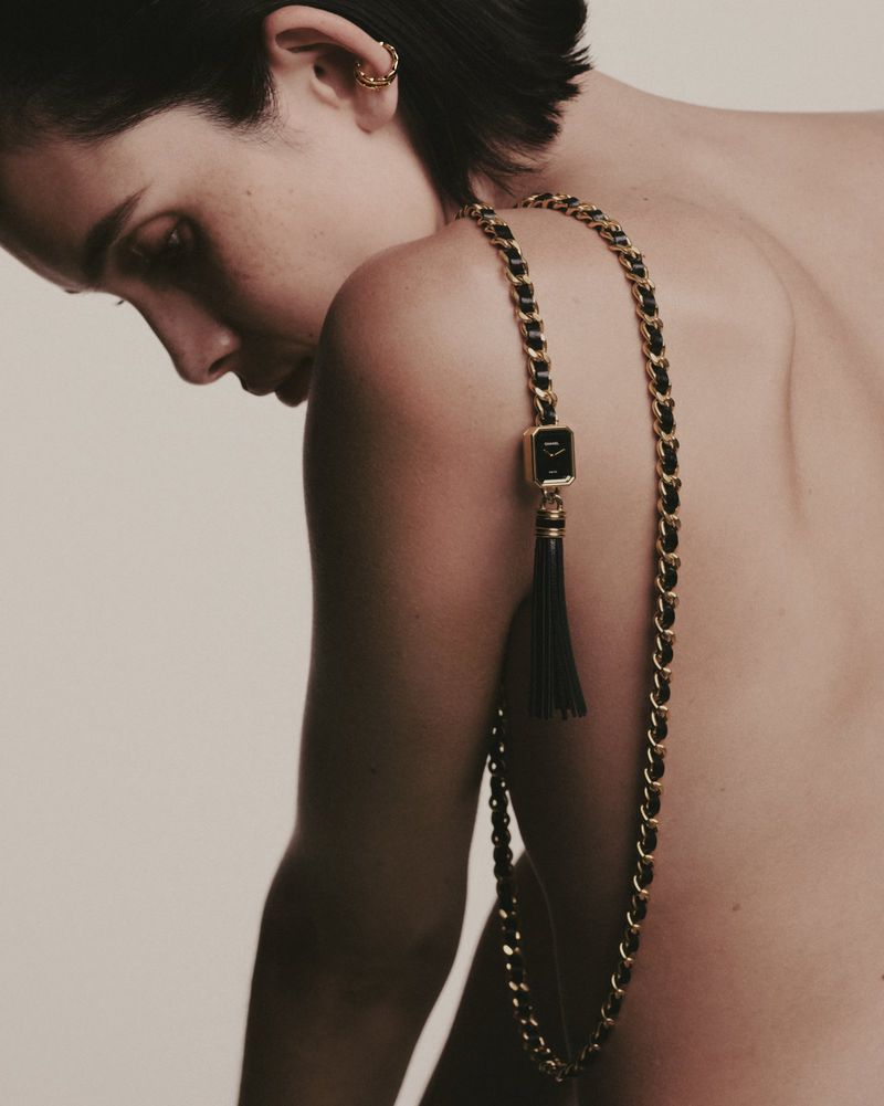 A nude adult female is visible in a close-up shot, adorned with a gold chain necklace. The backdrop is a clean, light neutral tone.