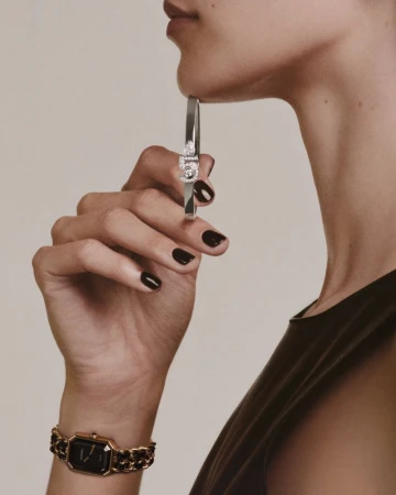 Close-up shot of a woman's wrist wearing a watch and holding up a silver bangle. The watch has a black face and gold chain band.