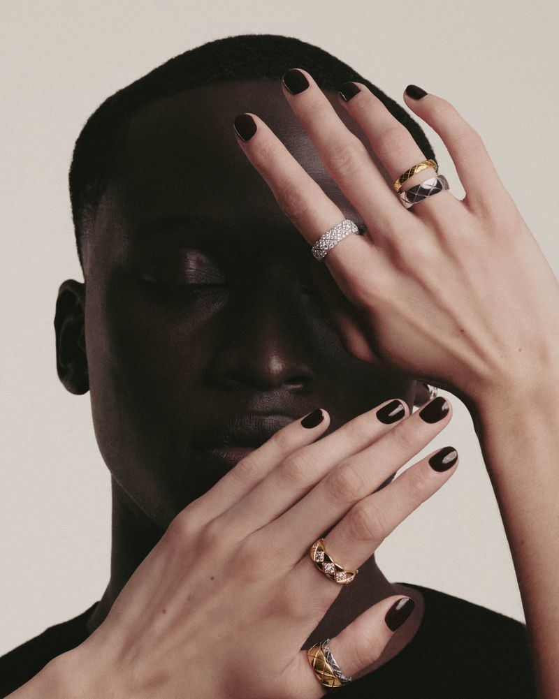 Close-up of Black male model showcasing rings; hands near face, dark nails, minimalist backdrop, beauty shot for jewelry advertising.