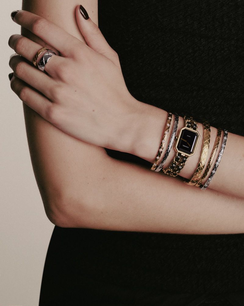 Close-up of a woman's arm adorned with a gold watch and a stack of bracelets and a ring against a neutral background.