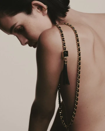 Model wearing a long gold chain necklace with a black and gold pendant draped over her shoulder against a neutral background.