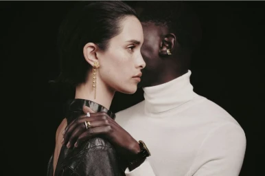Close-up profile portrait of a woman wearing gold earrings and a dark leather garment with a man standing behind her in a white turtleneck