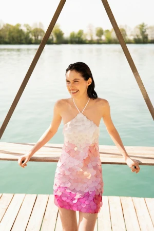 Smiling woman in pink ombre dress on wooden dock near lake. Summery fashion shoot in the sun with calm water and trees in background.