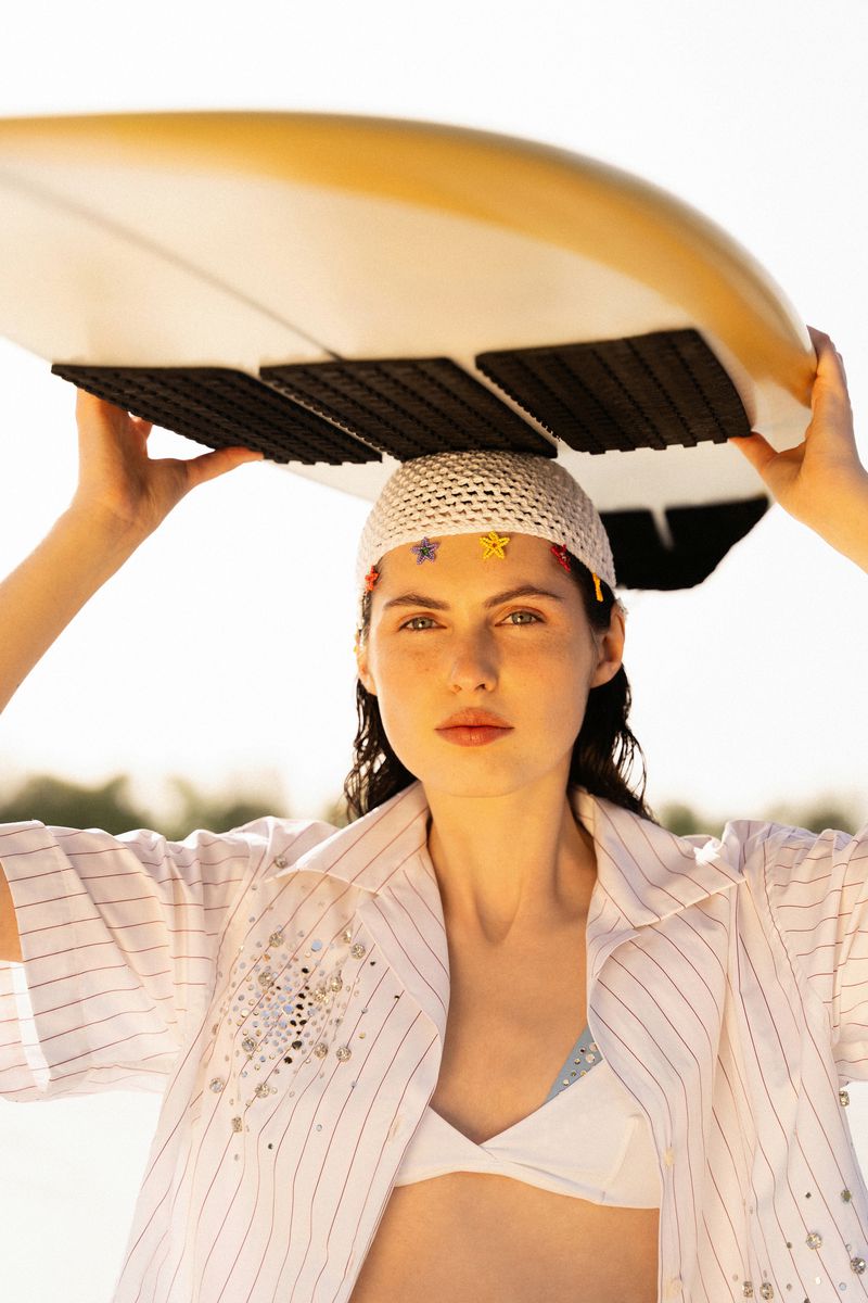 Brunette woman poses with a surfboard held over her head, wearing a striped shirt and white bikini top in an outdoor setting.