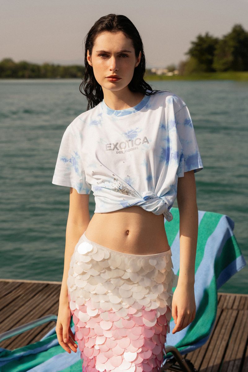Young woman in tie-dye top and sequined skirt stands on a dock by a lake in outdoor fashion portrait with lakeside background.