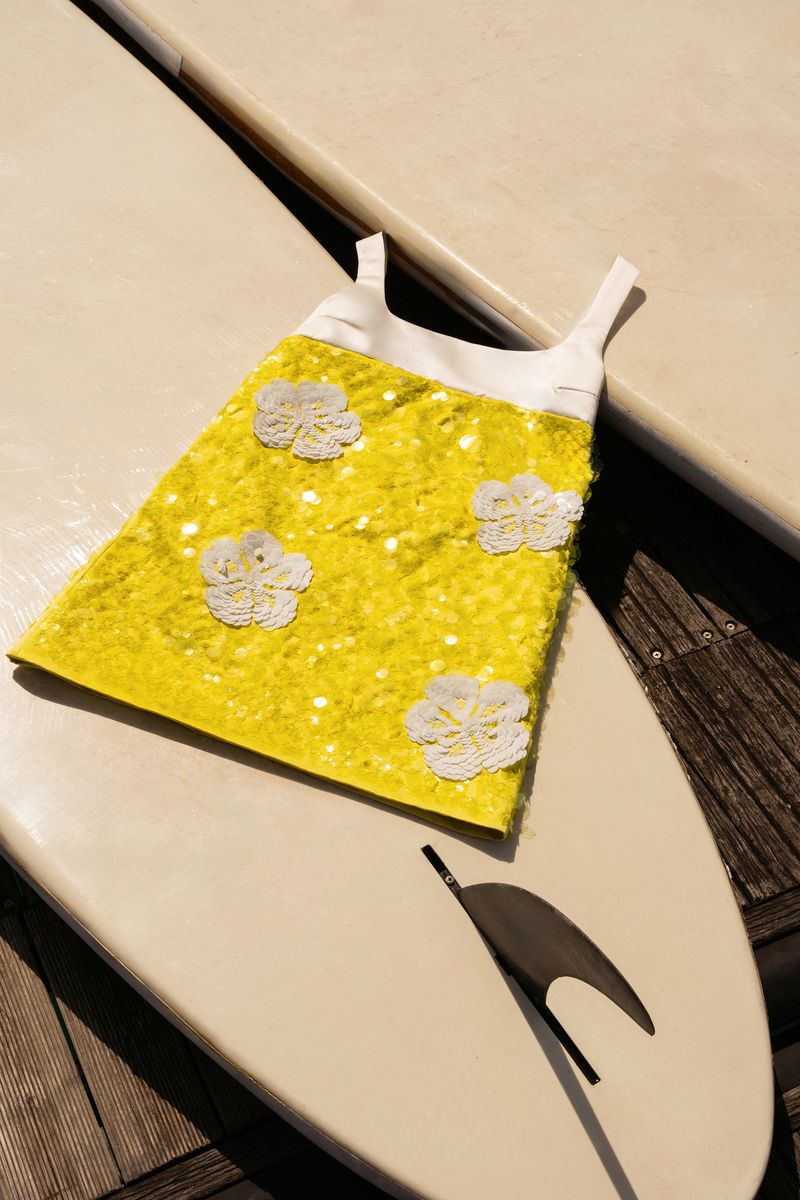 A yellow child's dress with white floral accents is displayed on a surfboard, with wood decking visible in the background.
