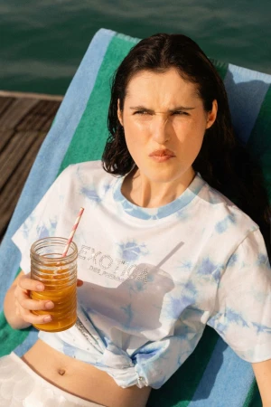 Woman relaxes on a striped beach towel, enjoying a refreshing orange drink in sunlight, wearing a tie-dye shirt by the water.