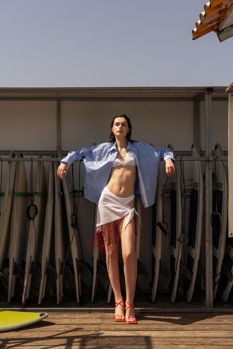 Woman in casual summer fashion posing on a deck with surfboards in background, wearing a bra top, wrap skirt, and oversized shirt.
