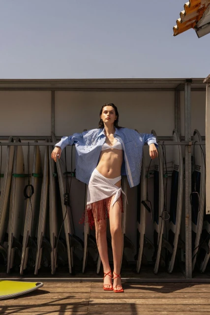 Woman in casual summer fashion posing on a deck with surfboards in background, wearing a bra top, wrap skirt, and oversized shirt.