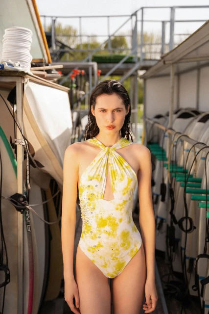 Woman in a yellow tie-dye swimsuit stands in front of surfboards, creating a sunny fashion portrait with a summer vibe.