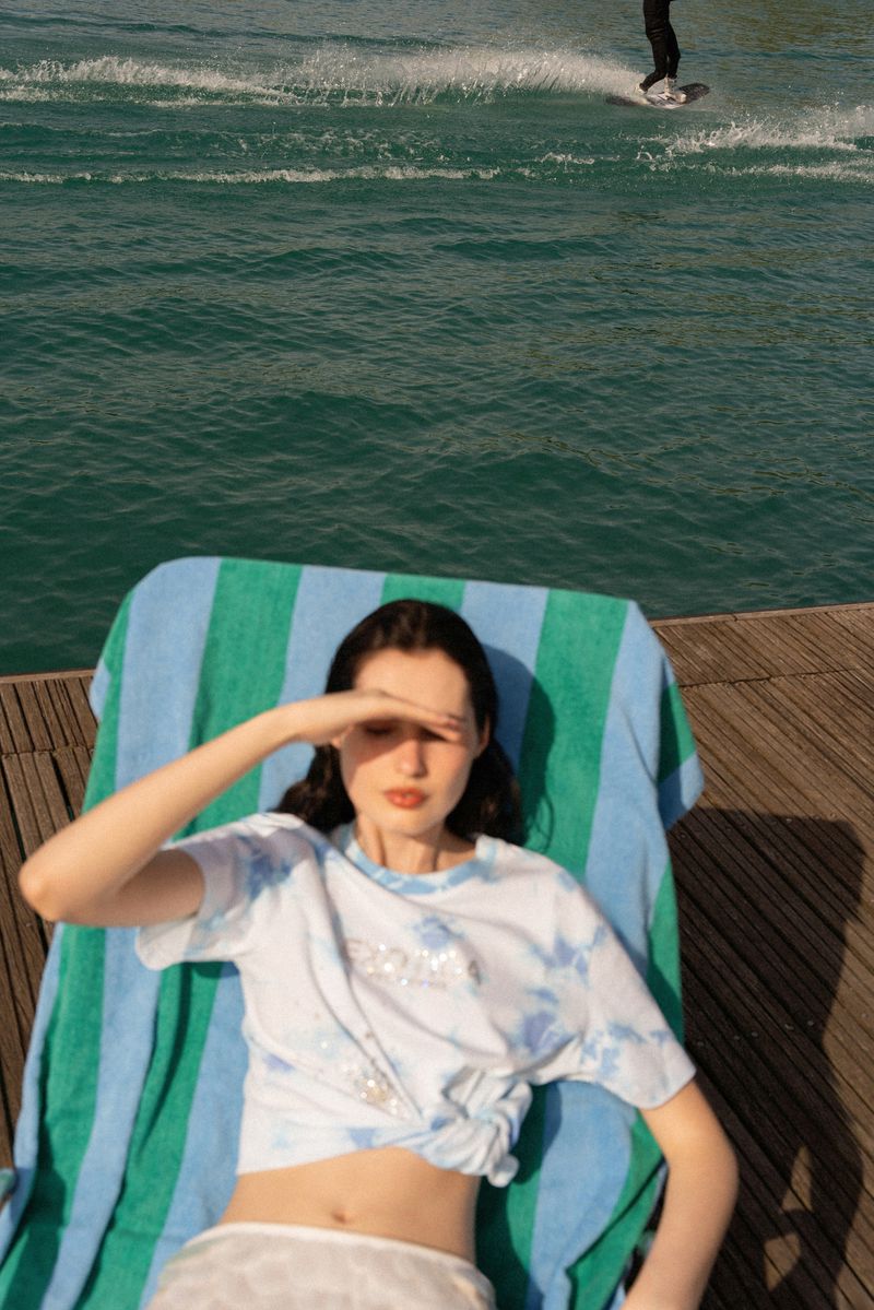 Adult female reclines on a striped towel at the water's edge, shielding eyes, with a distant person wakeboarding behind.