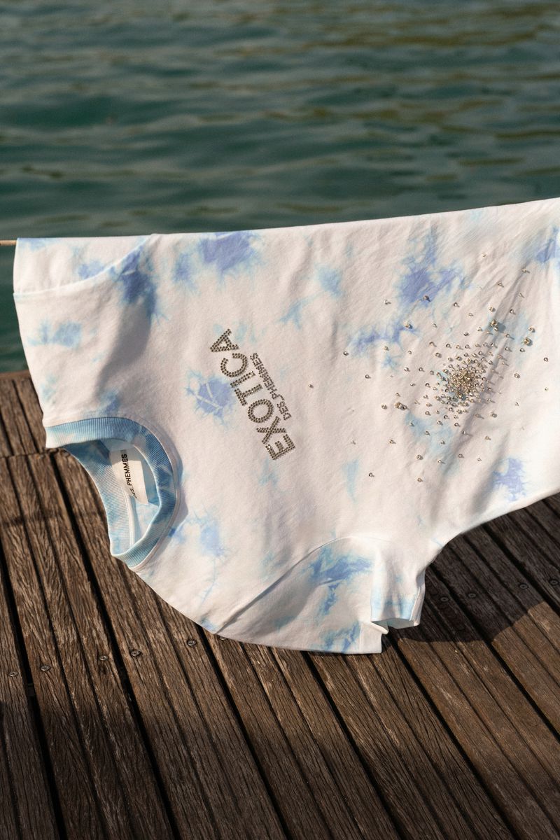 Hanging white t-shirt with blue dye pattern and rhinestones on wooden dock near rippling water, bathed in outdoor sunlight.