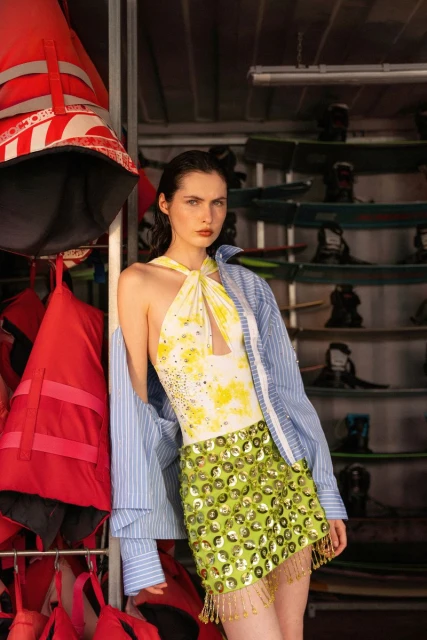 A woman in a yellow and white swimsuit and green skirt stands in a boat storage with life vests and wakeboards in the background.