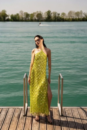 A fashionable woman poses in a lime green dress with a side slit on a wooden dock near a lake with a jet ski in the distance.