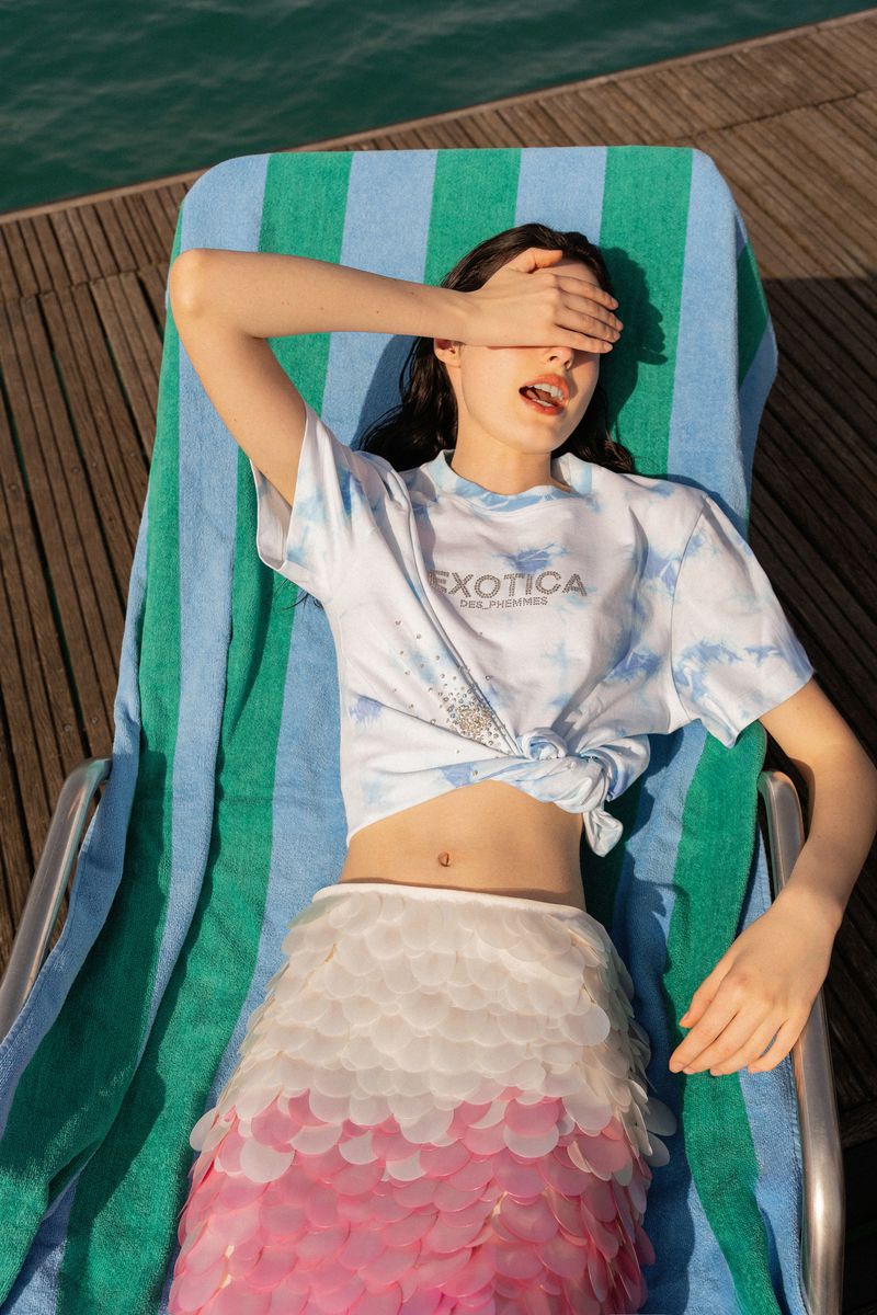 Young woman in a tie-dye t-shirt and petal skirt reclines on a striped towel and beach chair in the sun near turquoise water.
