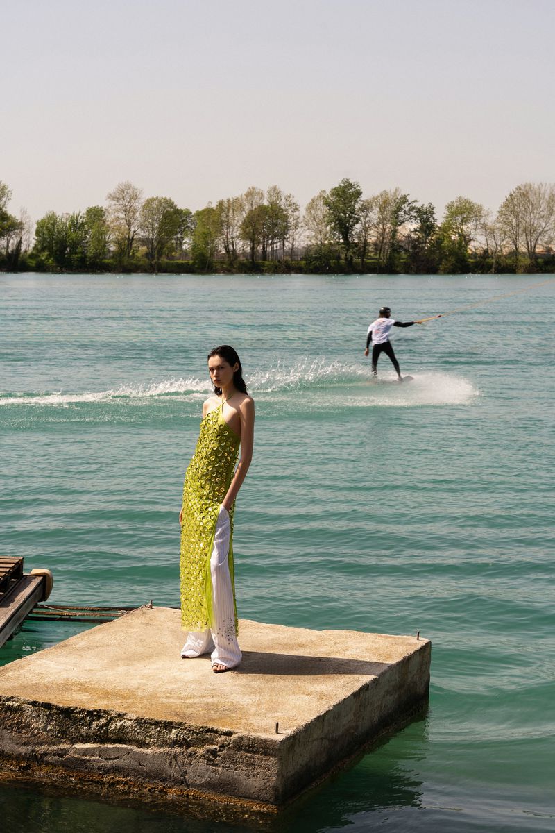 A woman in a yellow dress stands on a concrete pier next to the water, while a water skier glides across the river in the background.