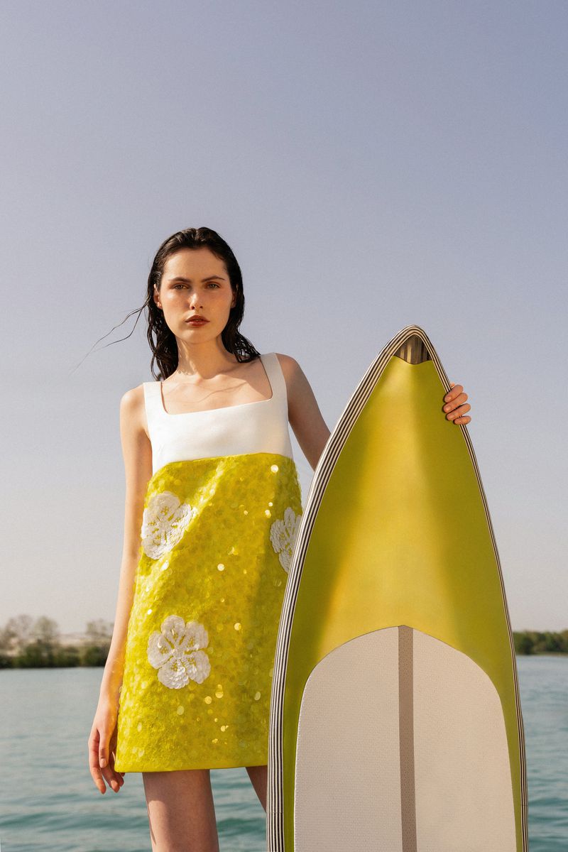 Brunette woman in a white and yellow dress holds a yellow surfboard in front of a blue sky and water background on a sunny day.