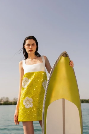 Brunette woman in a white and yellow dress holds a yellow surfboard in front of a blue sky and water background on a sunny day.