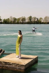 A model wearing a yellow embellished halter dress stands on a concrete dock by a body of water with a person wakeboarding in the distance.