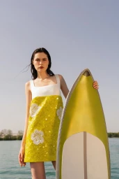 Model posing in a yellow sequined floral minidress while holding a chartreuse surfboard outdoors by the water.