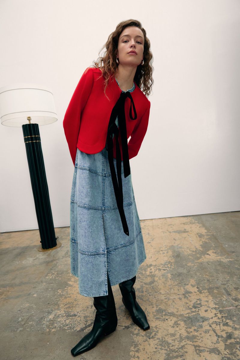 A slender woman poses in a red jacket, a denim skirt, and black boots against a white backdrop with a tall floor lamp.
