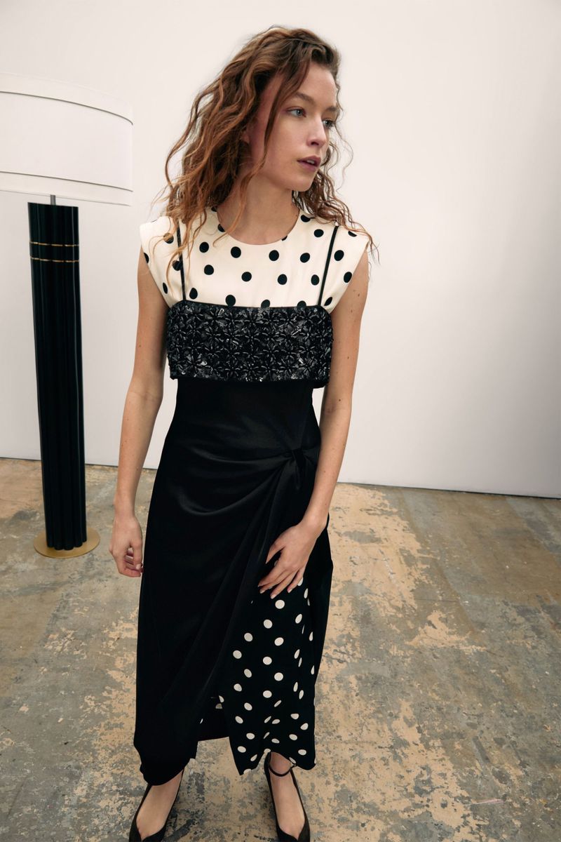 A slender woman with long, curly auburn hair poses in a studio wearing a sleeveless black and white polka dot dress with flats.