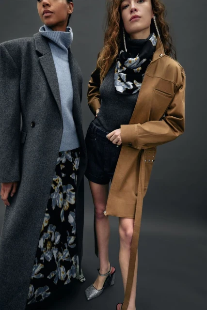 Two women pose in a fashion studio wearing stylish coats and dresses. One is wearing a grey wool coat with a light blue sweater.