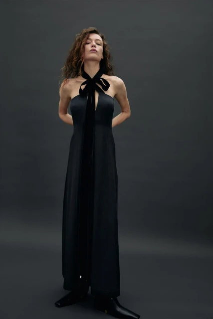 Elegant woman in black halter dress with velvet bow poses in a studio with a dark background, showcasing fashion and style.