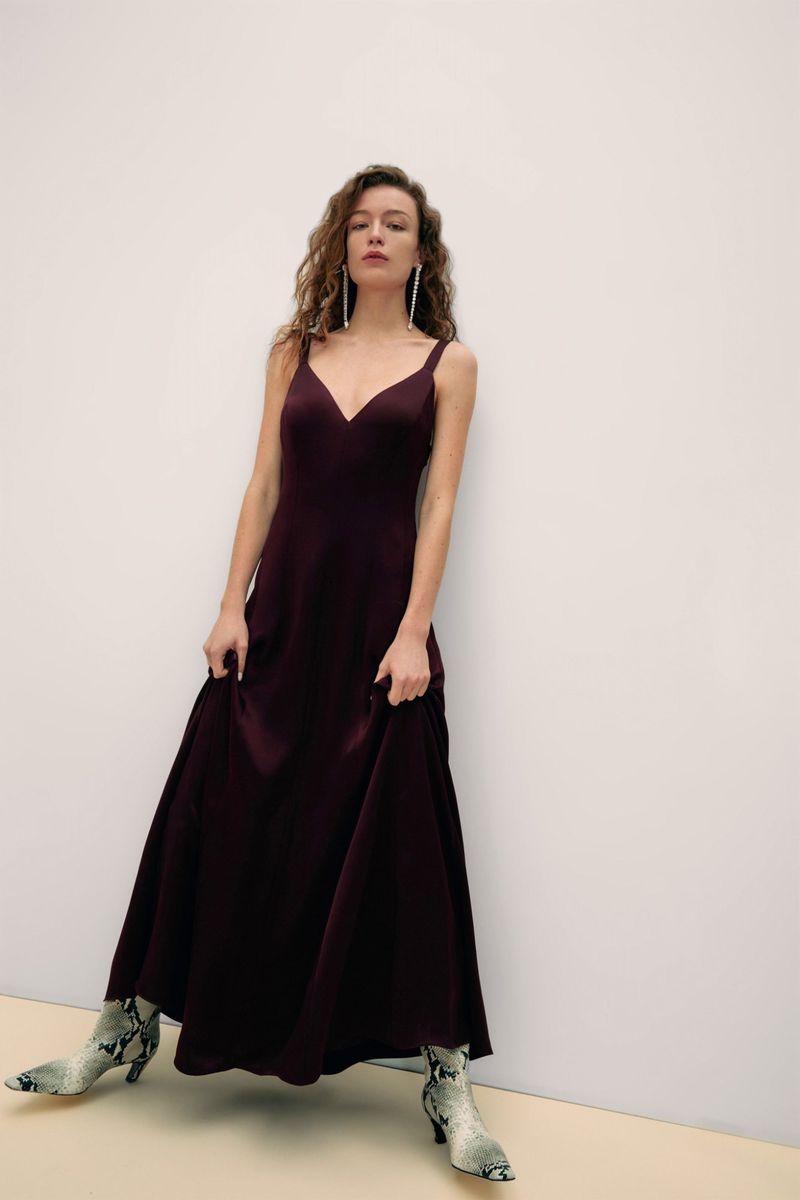 Elegant woman poses in a dark plum dress, holding the skirt, and wearing snakeskin print boots in a studio setting with a cream backdrop.