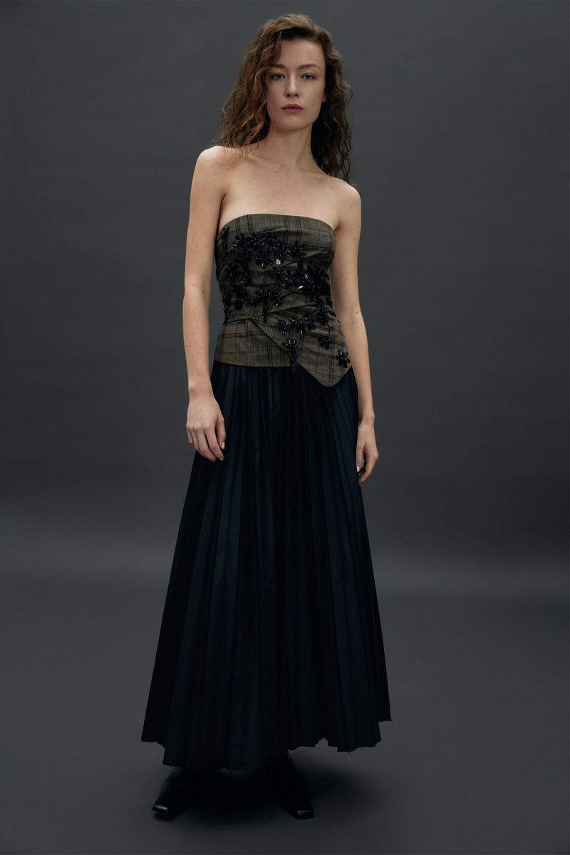 Model Hazel Jane wearing a strapless plaid embellished top and a black pleated maxi skirt against a dark grey seamless studio backdrop.