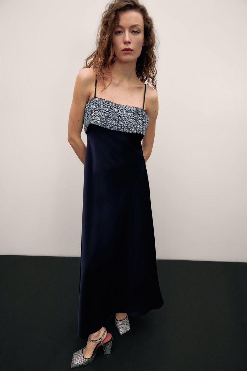 Elegant woman posing in a sleeveless, floor-length navy dress with a sequined bodice and glittery high heels in a bright studio.