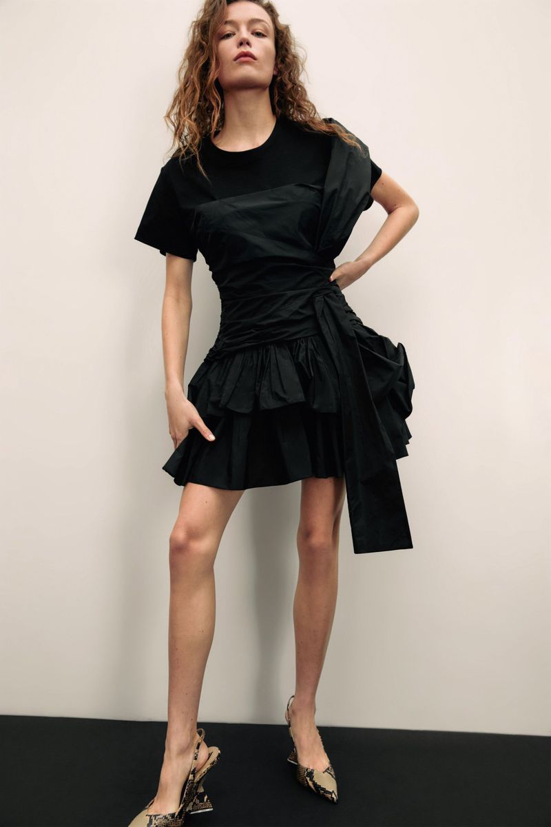 A fair-skinned woman with curly brown hair poses in a studio wearing a black dress and patterned heels, against a neutral backdrop.