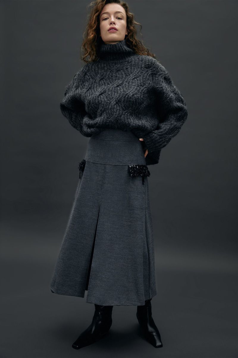 Elegant woman in grey sweater and midi skirt poses against a solid grey background in a fashion studio setting.