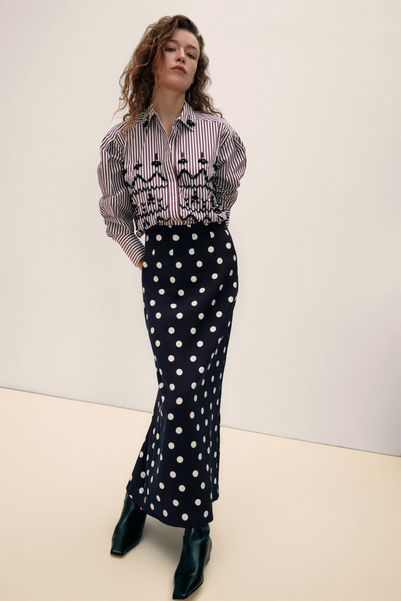Studio shot of a woman wearing a striped shirt and polka dot skirt, posing indoors against a neutral background, fashion editorial.