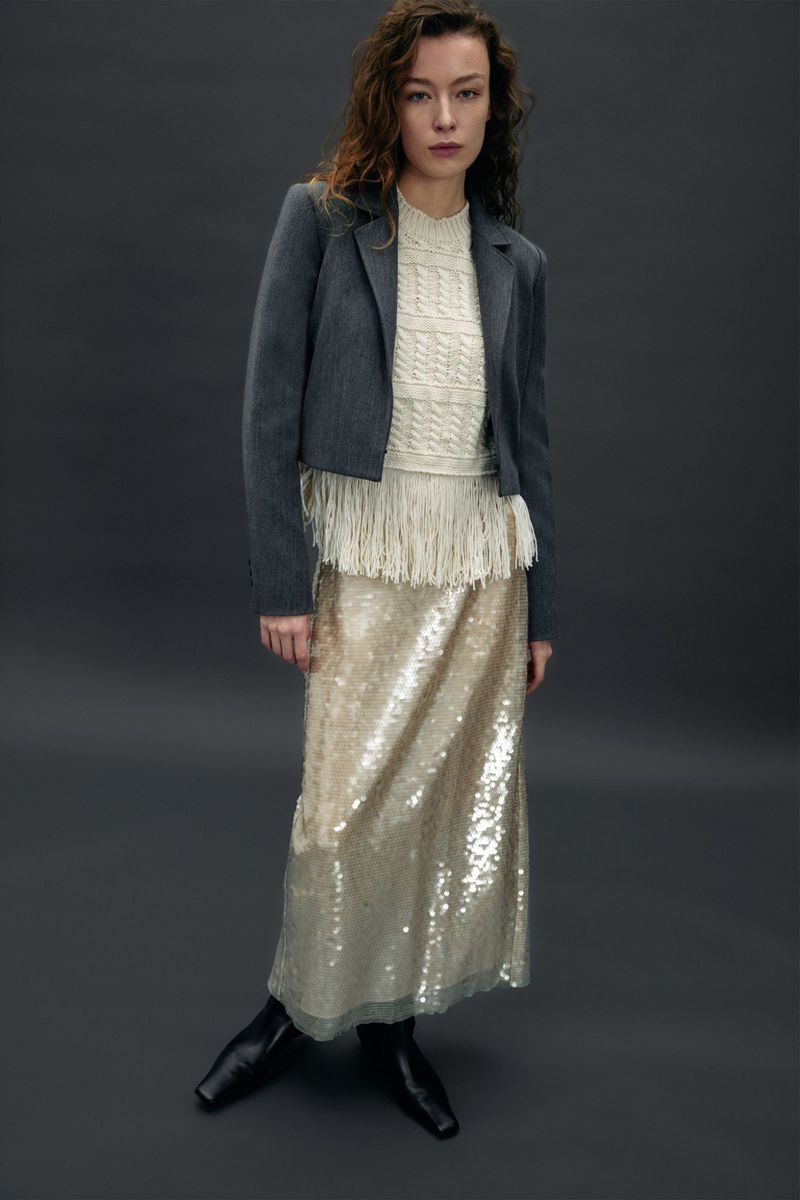 Fashionable woman in a grey jacket, fringe top, and sequin skirt poses in a studio, showcasing a modern and stylish outfit.