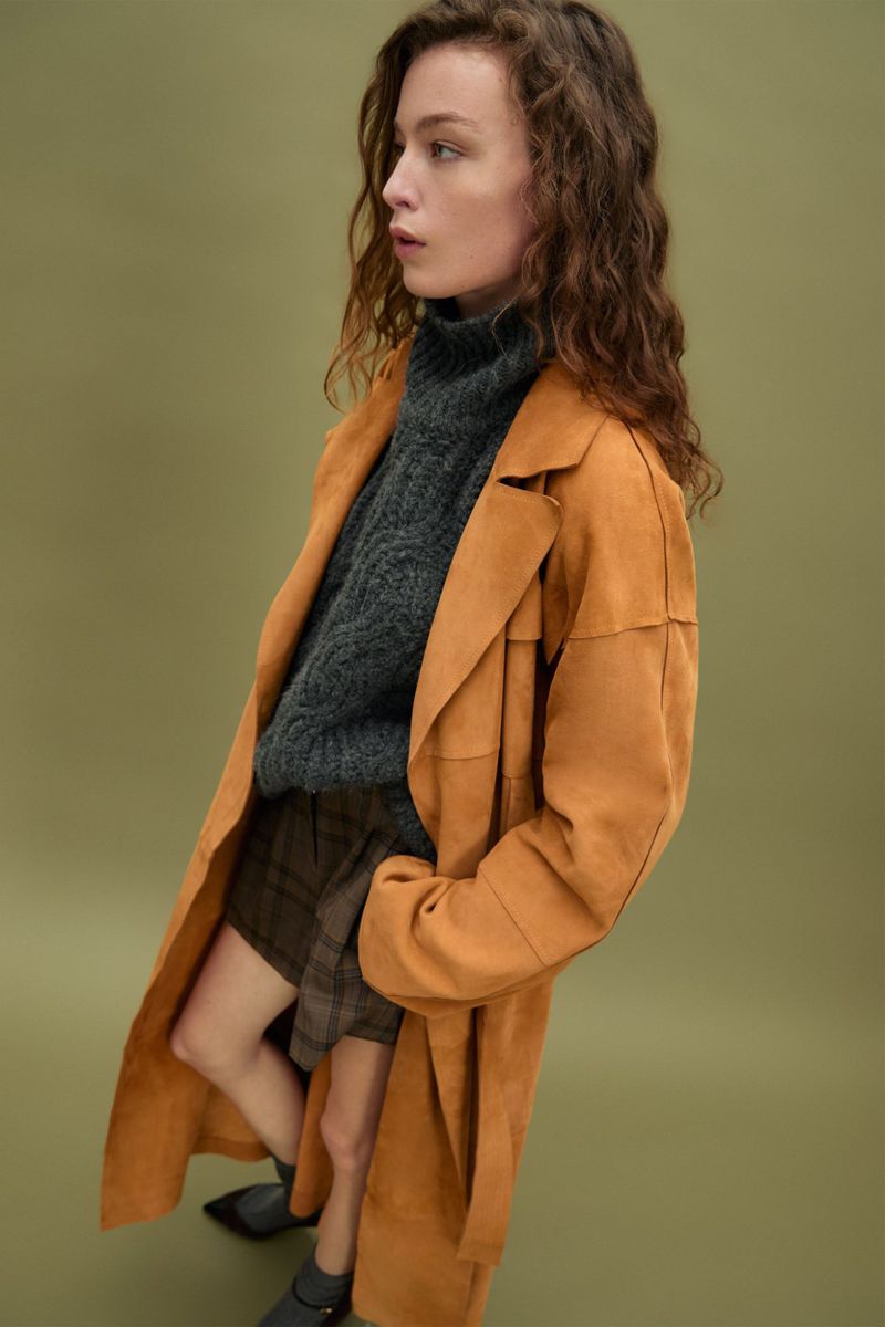 An adult woman poses wearing an orange coat, gray turtleneck sweater, and plaid skirt in a studio with a green background.