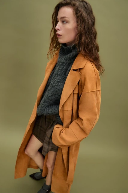 An adult woman poses wearing an orange coat, gray turtleneck sweater, and plaid skirt in a studio with a green background.