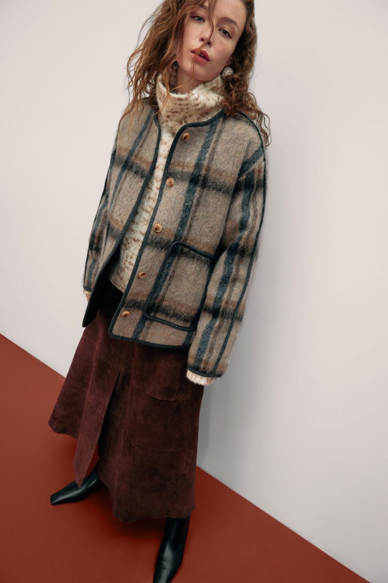 A woman in a beige plaid jacket and brown skirt stands against a neutral backdrop with a two-toned rust and white floor.