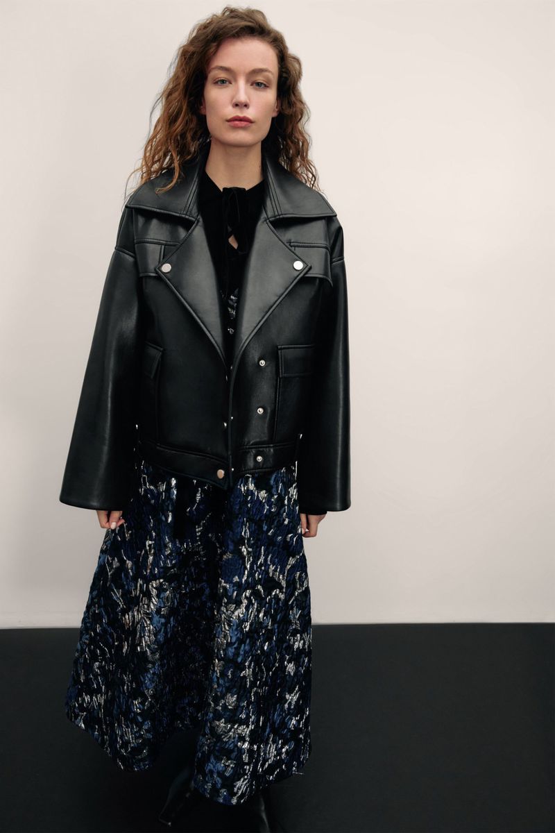 A fashionable woman in a black leather jacket and patterned midi skirt poses in a photo studio with neutral walls and dark floor.