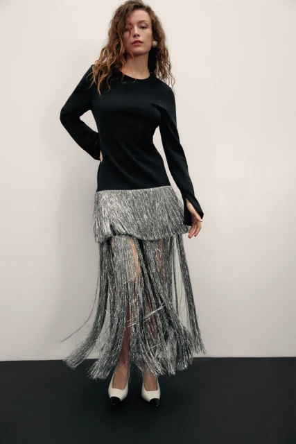 Woman in a black long-sleeved dress with silver fringe poses indoors. The dress is paired with cream and black heels against a neutral background.