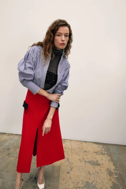 Adult woman in a photo studio wearing a striped shirt, red skirt, and white high heels posing against a neutral colored wall.