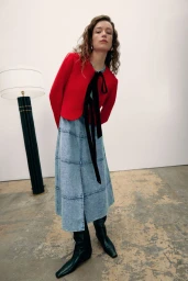 Model Hazel Jane wearing a red cropped cardigan with black tie, tiered denim skirt, and black boots against a white studio wall.