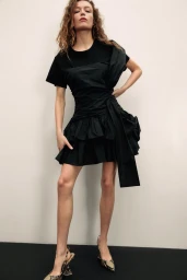 Model wearing a black dress with ruffled skirt and animal-print heels against a neutral studio backdrop